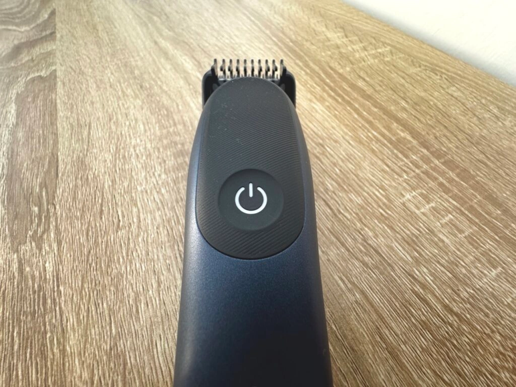 Ein kompletter Braun-Rasierer liegt quer auf einem Holztisch. Man erkennt das schlanke Design mit einem großen Einschaltknopf in der Mitte. Das Gerät ist schwarz-blau und wirkt ergonomisch geformt.