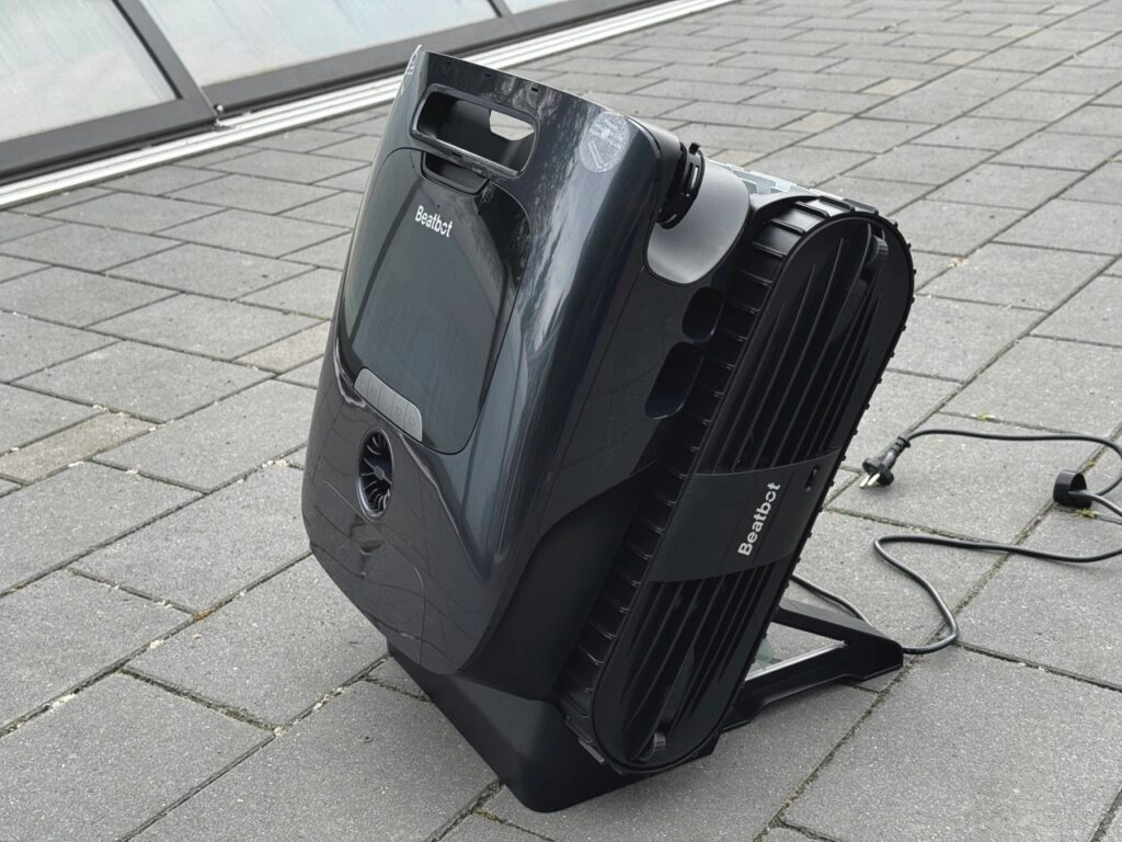 Der Roboter steht senkrecht in seiner schwarzen Ladestation. Sichtbar sind die Lüftungsschlitze an der Seite und das Stromkabel, das am Boden entlang verläuft.