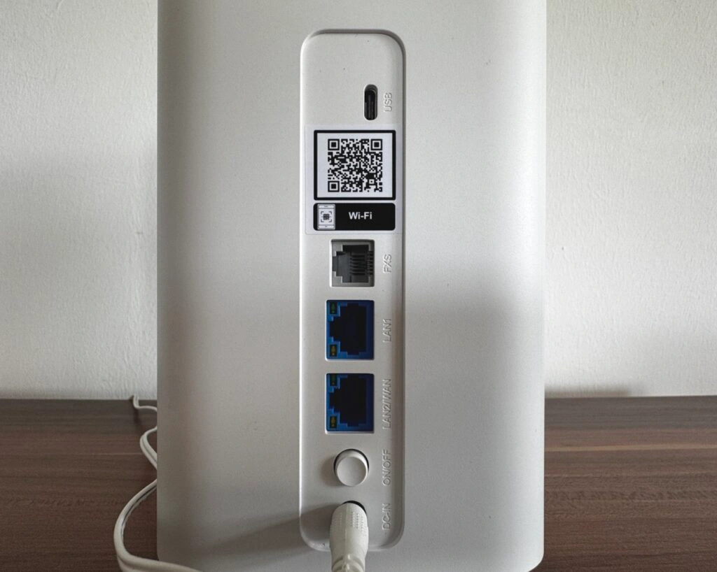 Nahaufnahme der Rückseite mit einem USB-Anschluss, einem FXS-Port, zwei blauen LAN-Ports, Ein-/Ausschalter, Stromanschluss und QR-Code für WLAN-Zugang.