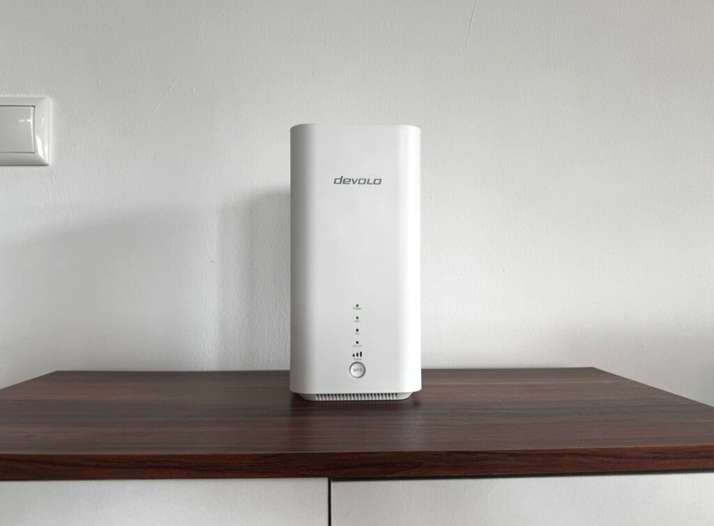 Der devolo-Router auf einem weißen Möbelstück mit dunkler Holzplatte vor einer glatten Wand.