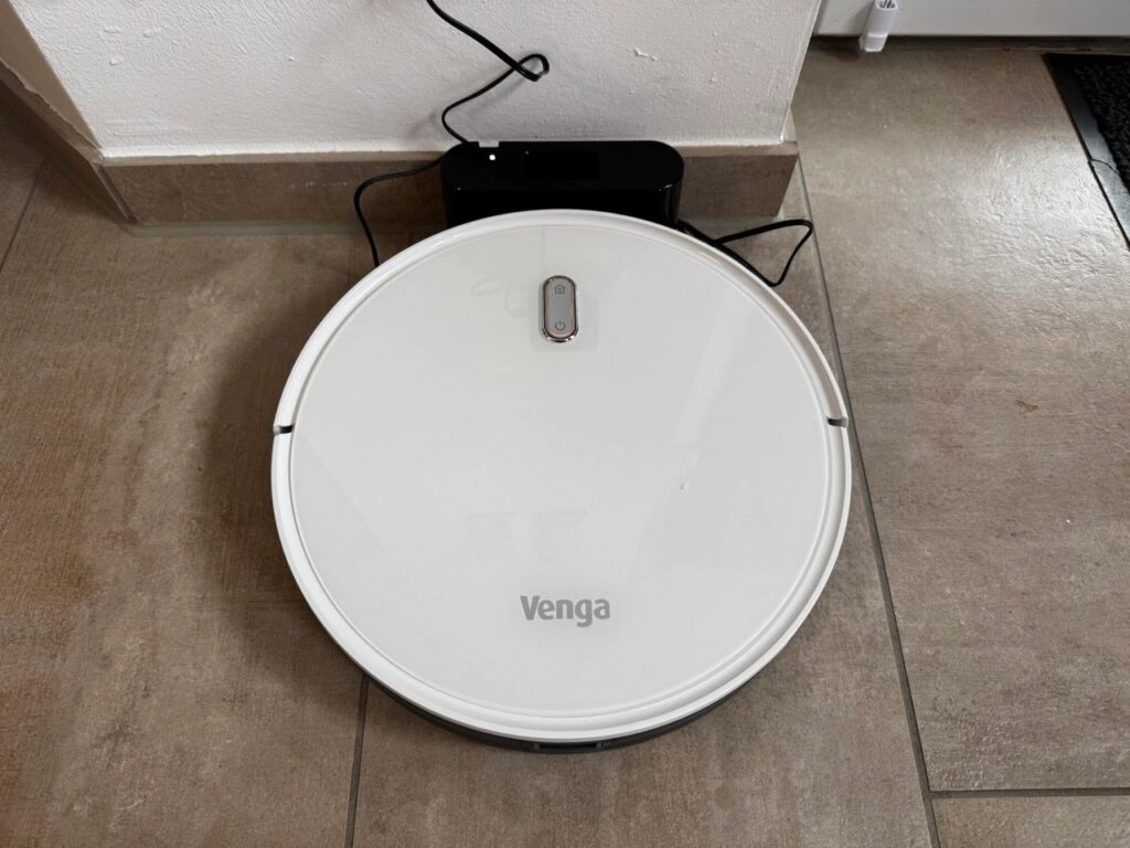 Ein weißer Saugroboter mit der Aufschrift „Venga“ befindet sich an seiner schwarzen Ladestation. Das Gerät lädt, das Design ist schlicht und rund.