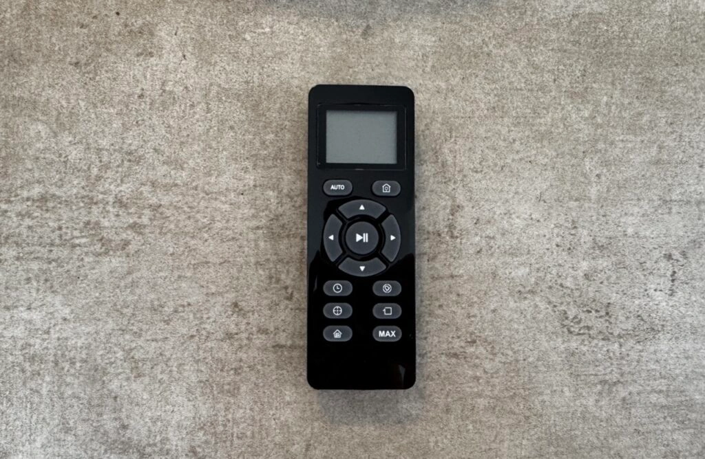 Eine schwarze Fernbedienung mit Display liegt auf einem grauen Boden. Zu sehen sind Richtungstasten, Auto-Modus, Start/Stopp und weitere Funktionstasten wie „Max“ oder „Home“.