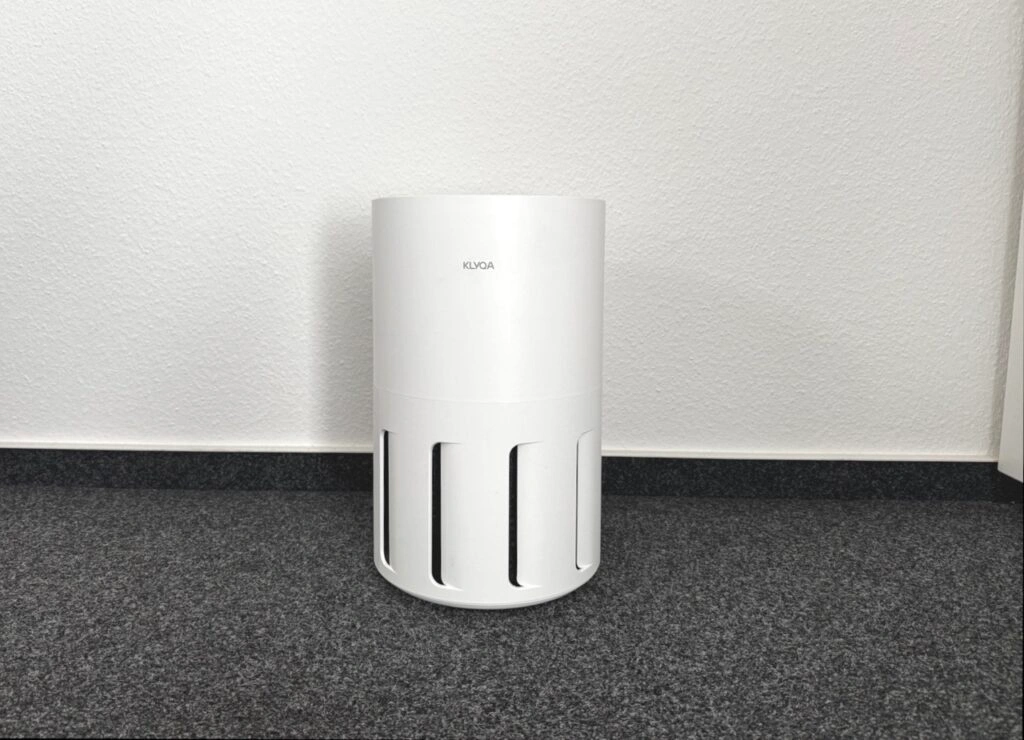 Ein weißer, zylindrischer Luftreiniger steht auf Teppichboden an einer weißen Wand. Das Gerät trägt das Logo „KLYQA“ und zeigt keine sichtbaren Tasten oder Anzeigen.