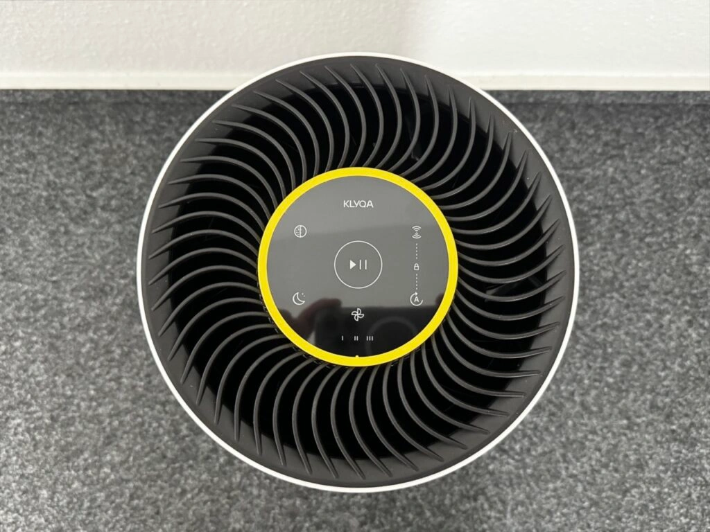 Draufsicht auf die Oberseite des Luftreinigers mit schwarzem Lüftergitter und zentralem Bedienfeld mit mehreren Touch-Symbolen. Ein gelber Ring umrandet das Panel.