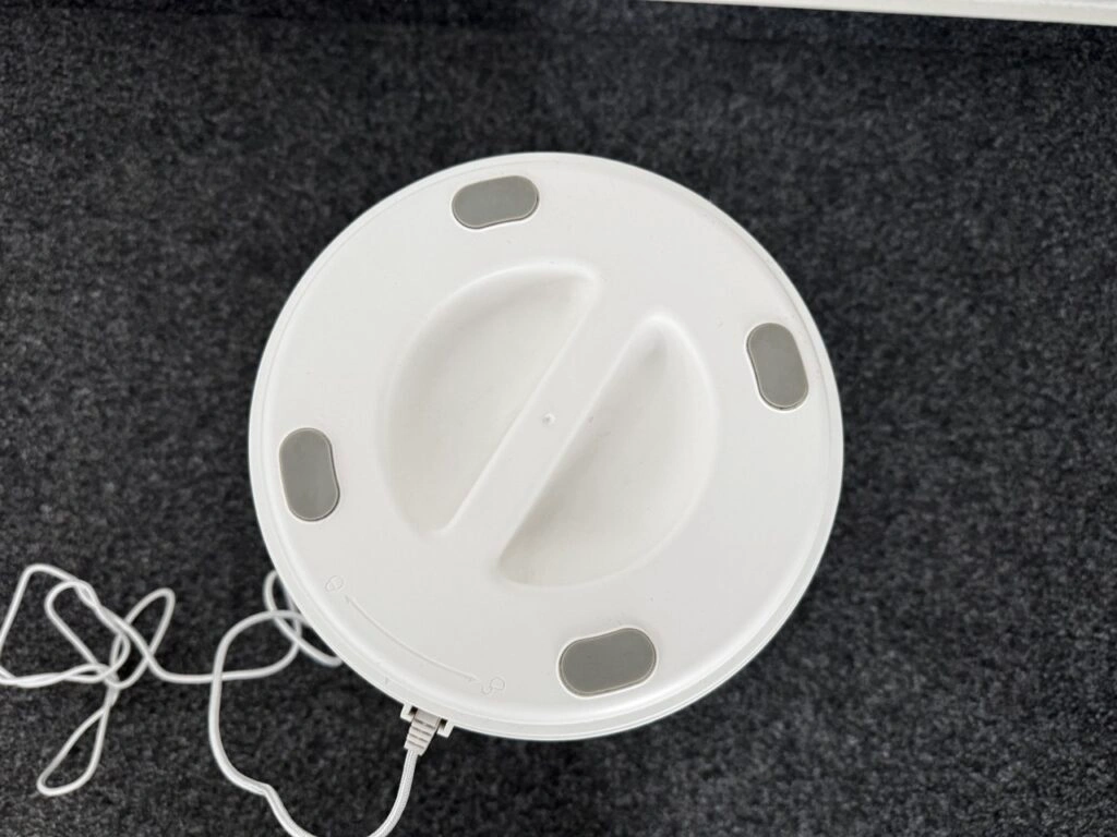 Ansicht der Geräteunterseite mit vier rutschfesten Füßen und einer Drehmarkierung zum Öffnen. Ein Kabel führt vom Gerät weg.