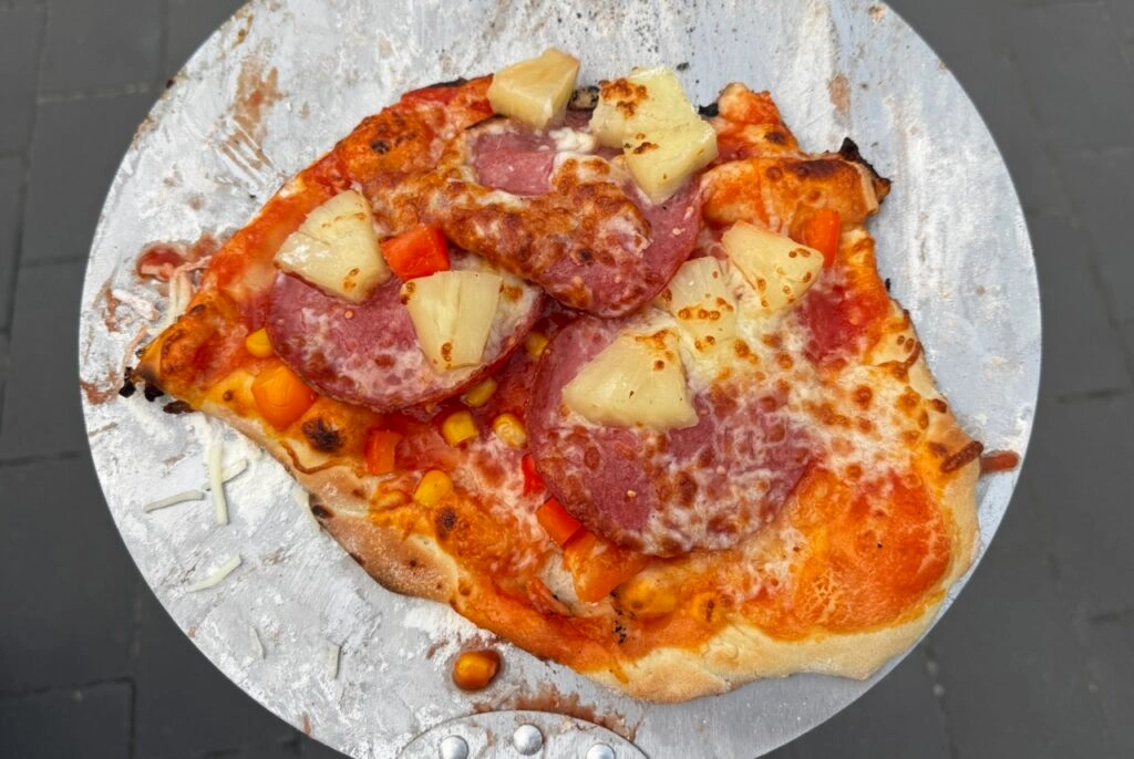 Pizza Hawaii mit Käse, Ananas, Salami, Mais und Paprika auf einem Pizzaheber.