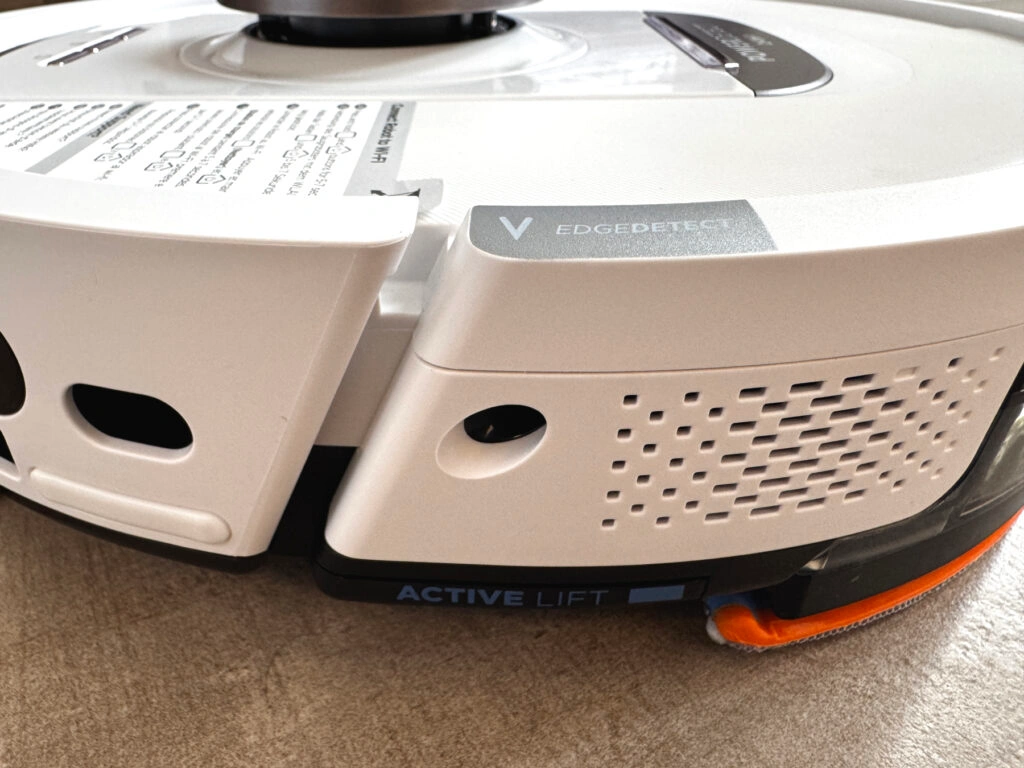 Nahaufnahme der Seitenansicht des Shark PowerDetect NeverTouch Pro 2-in-1. Sichtbar sind die EdgeDetect- und Active Lift-Funktionen. Die EdgeDetect-Technologie soll Schmutz aus Ecken effizienter entfernen, während Active Lift das Hinterteil des Roboters anhebt, um Teppiche beim Wischen trocken zu halten. Das Wischpad ist teilweise sichtbar, mit orangefarbenem Rand.